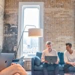 Three men seated on couches, engaged with laptops, sharing insights on organizational knowledge and collaboration.