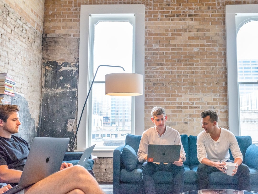 Three men seated on couches, engaged with laptops, sharing insights on organizational knowledge and collaboration.